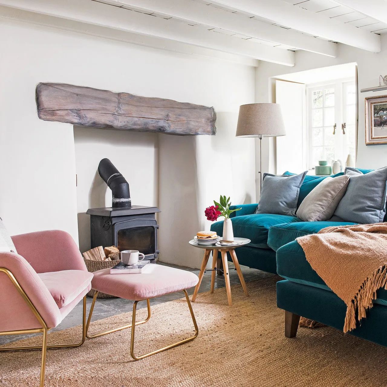 Thick timber mantel above a stove beside a pink chair and footstool opposite a deep teal sofa