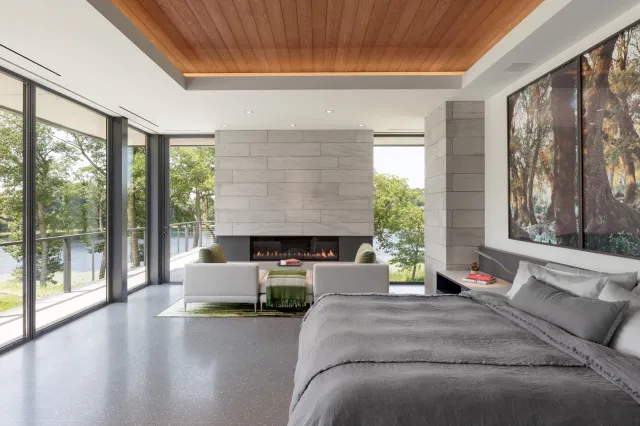A quiet bed sits below a timber ceiling beside broad windows and pale architectural surfaces.