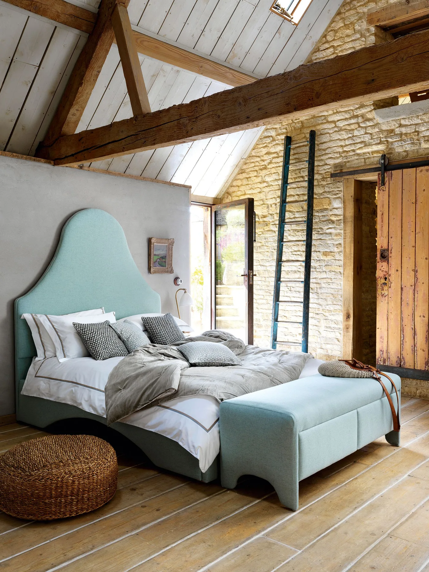 Bedroom with a tall pale blue upholstered headboard and matching bench, exposed stone wall, wooden roof beams and a sliding wooden door.