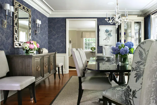 Deep blue patterned walls above white paneling beside a timber sideboard and pale dining chairs