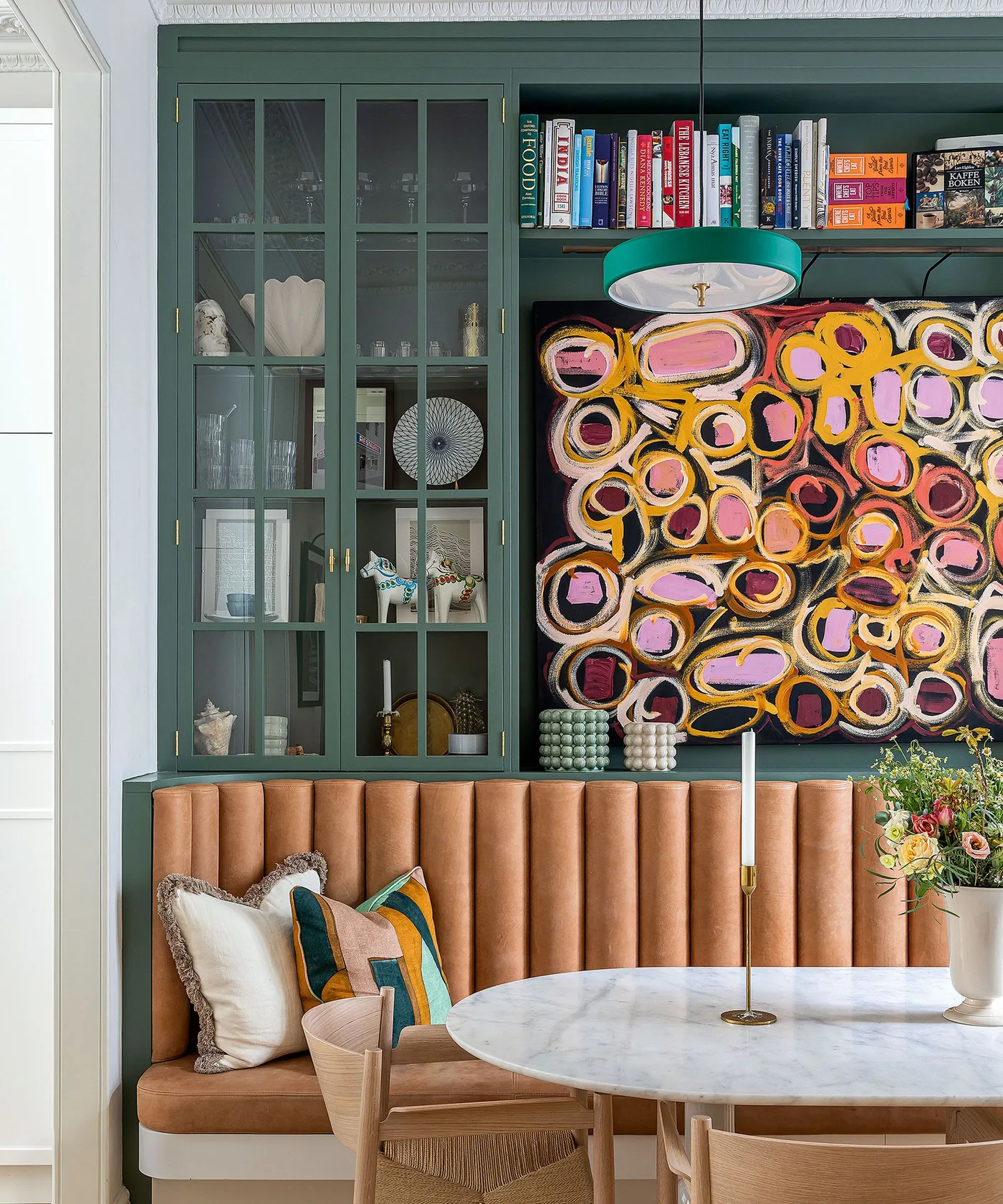 Dark green glazed cupboards and a book shelf above a tan upholstered dining bench with a colorful patterned panel