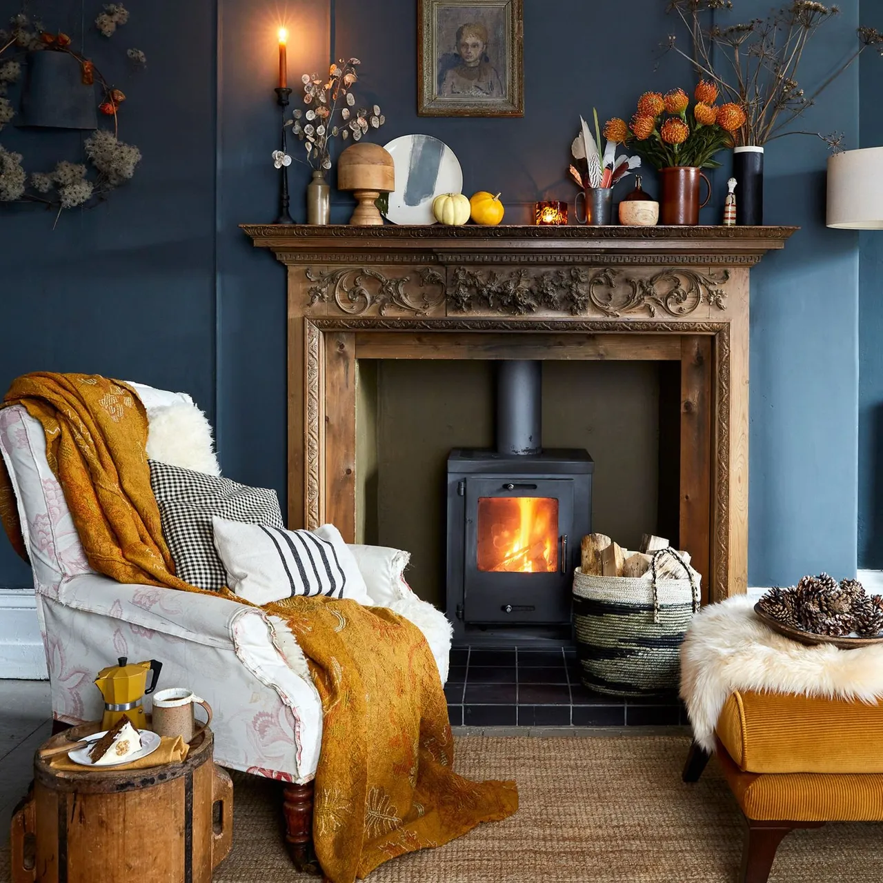 Carved wood mantel against dark blue wall with small pumpkins, orange flowers, stove and mustard textiles