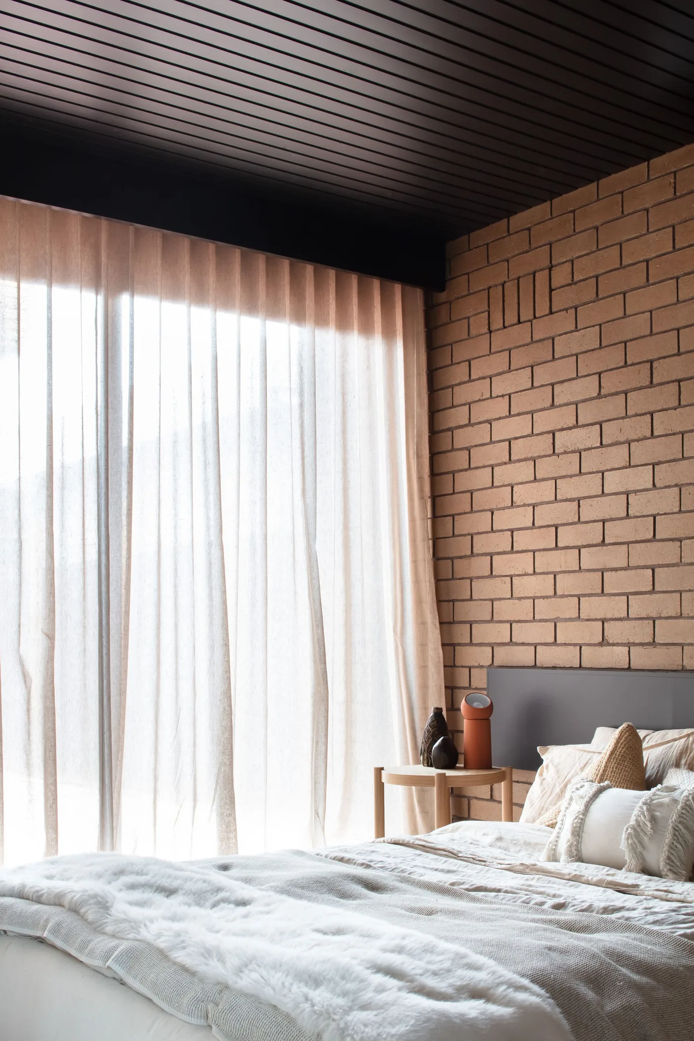 Warm brick bedroom wall beneath a dark lined ceiling beside pale curtains and light bedding