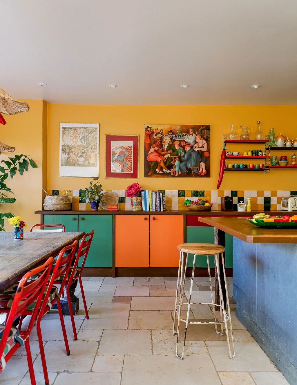 Orange and green cabinet doors sit beneath yellow walls with framed artwork, colorful shelves and a mixed square-tile backsplash beside red dining chairs