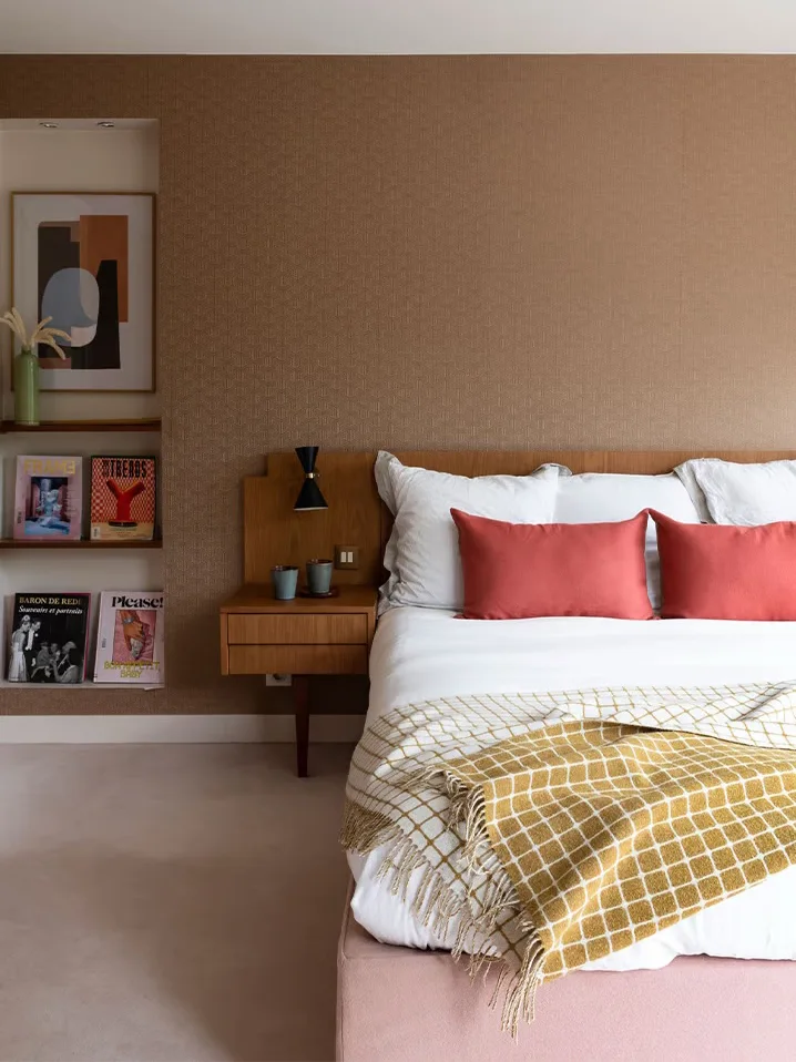 Small bedroom shelves hold books beside a wood headboard and cabinet below textured brown walls with pale bedding, coral cushions and a patterned yellow throw