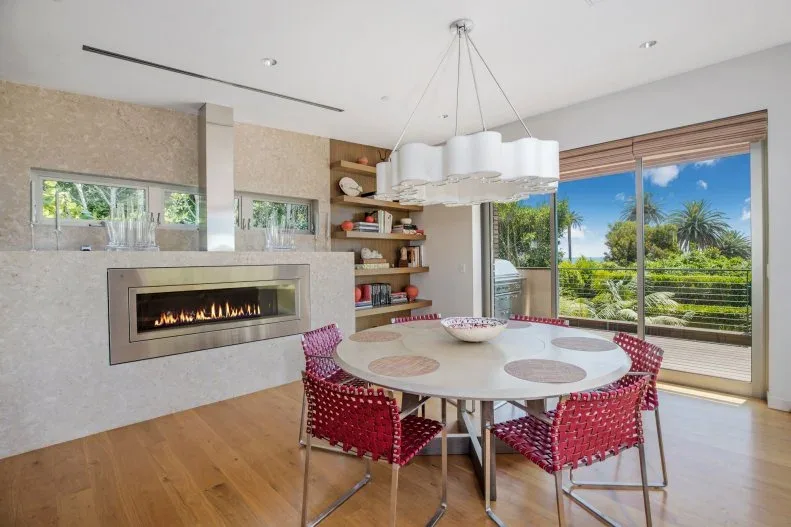Grouped pale cylindrical shades suspended from a shared ceiling point above a round pale table and red woven chairs, beside a linear fireplace and broad window.
