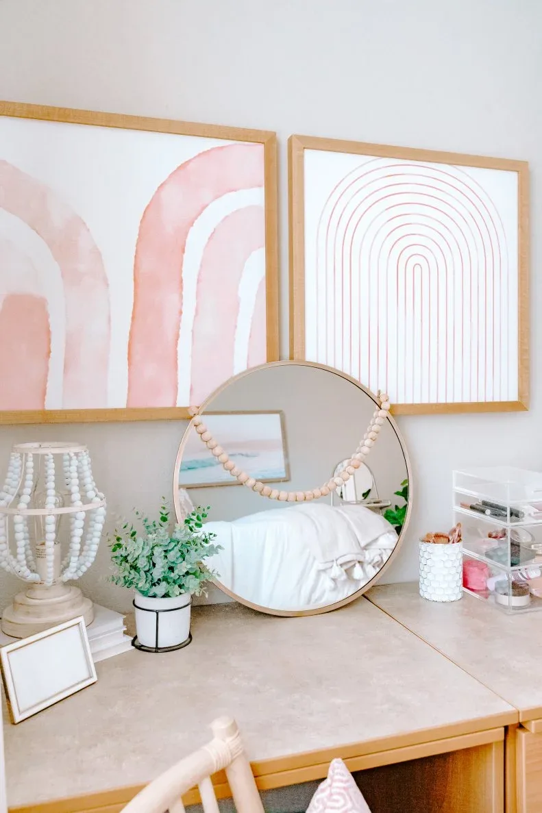 Wooden desk surface with round mirror, pale framed prints, small plant, pen pot and clear stacked organizers.