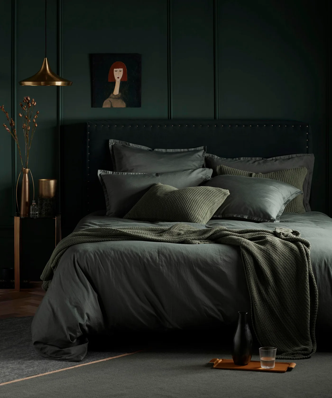 Dark green paneled bedroom with a dark upholstered headboard, green bedding, gold-colored pendant, and portrait above the bed.
