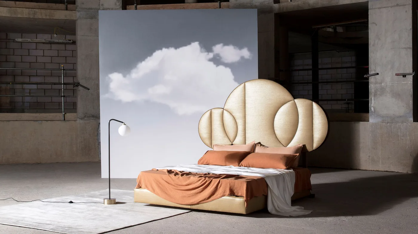 Staged bed with a pale rounded segmented moon headboard, rust bedding and a cloud backdrop within a gray interior.
