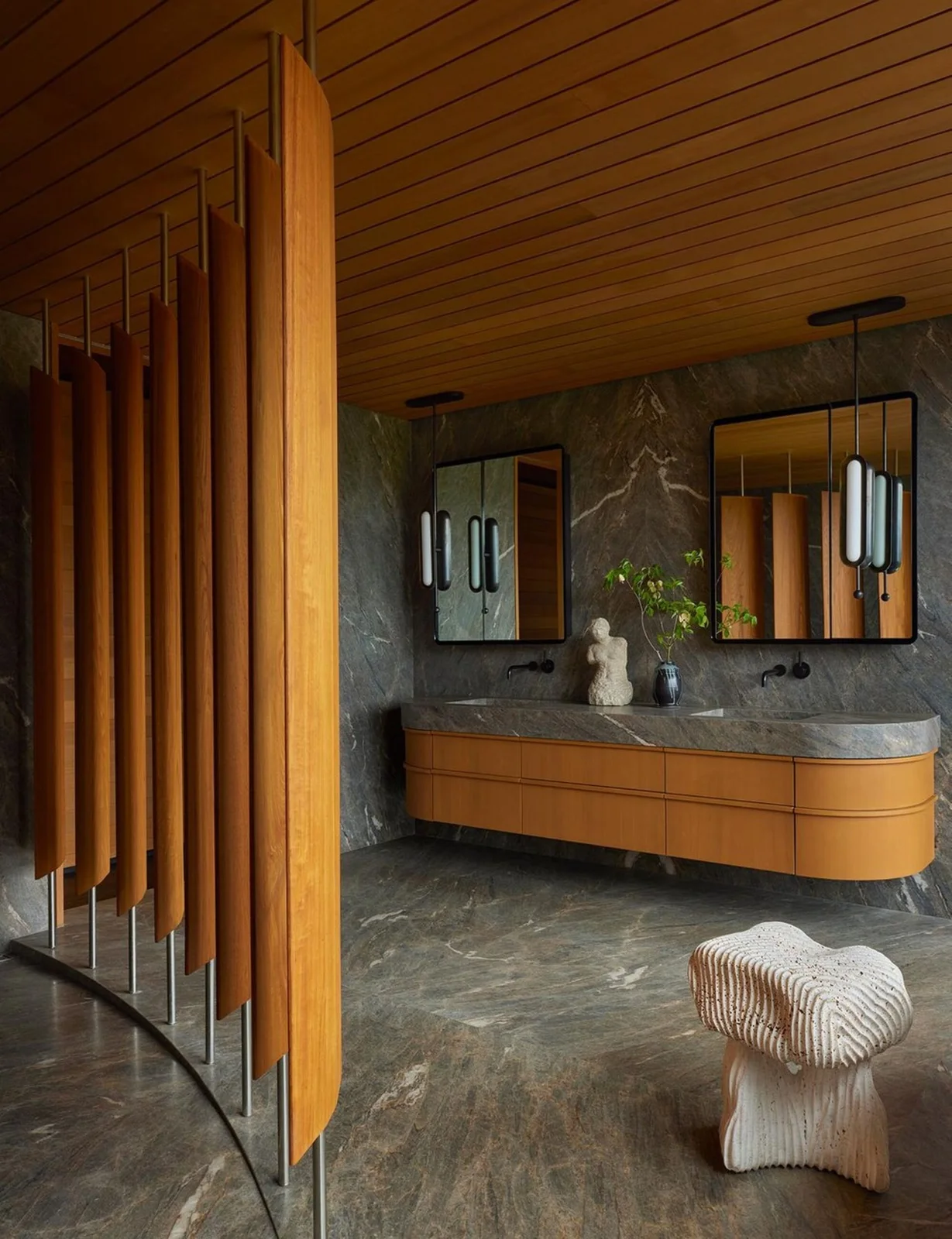 A curved wood bathroom vanity stands beneath two framed mirrors beside gray veined surfaces, a wood ceiling, vertical wood screens and a sculptural stool