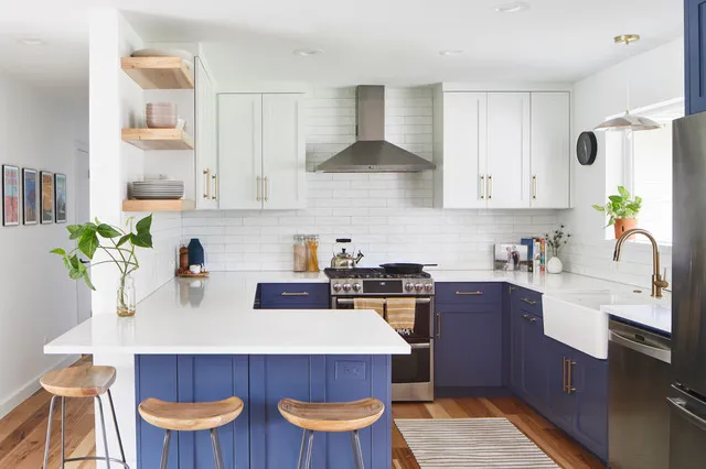 Navy lower cabinets and a small island sit beneath white upper cupboards and a simple metallic hood beside wooden stools and pale wall tiles