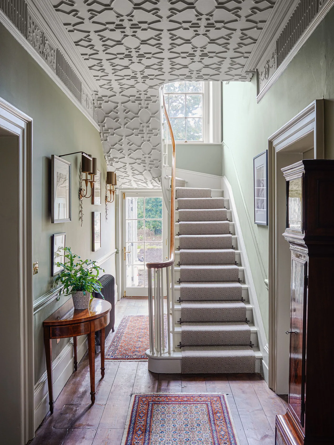 Traditional hallway with a patterned ceiling, pale stair runner, wooden handrail and small rugs on the floor.