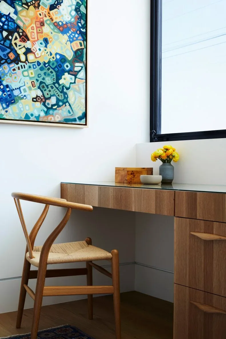 Home office detail with wooden drawer desk, wooden chair with woven seat, colorful abstract artwork, pale wall and tall dark-framed window.