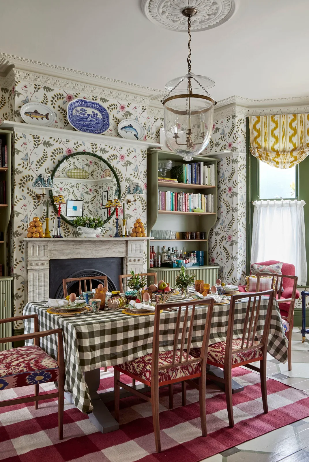 Leafy wallpaper surrounds shelves, decorative plates and an oval mirror above a checked tablecloth and a red checked rug