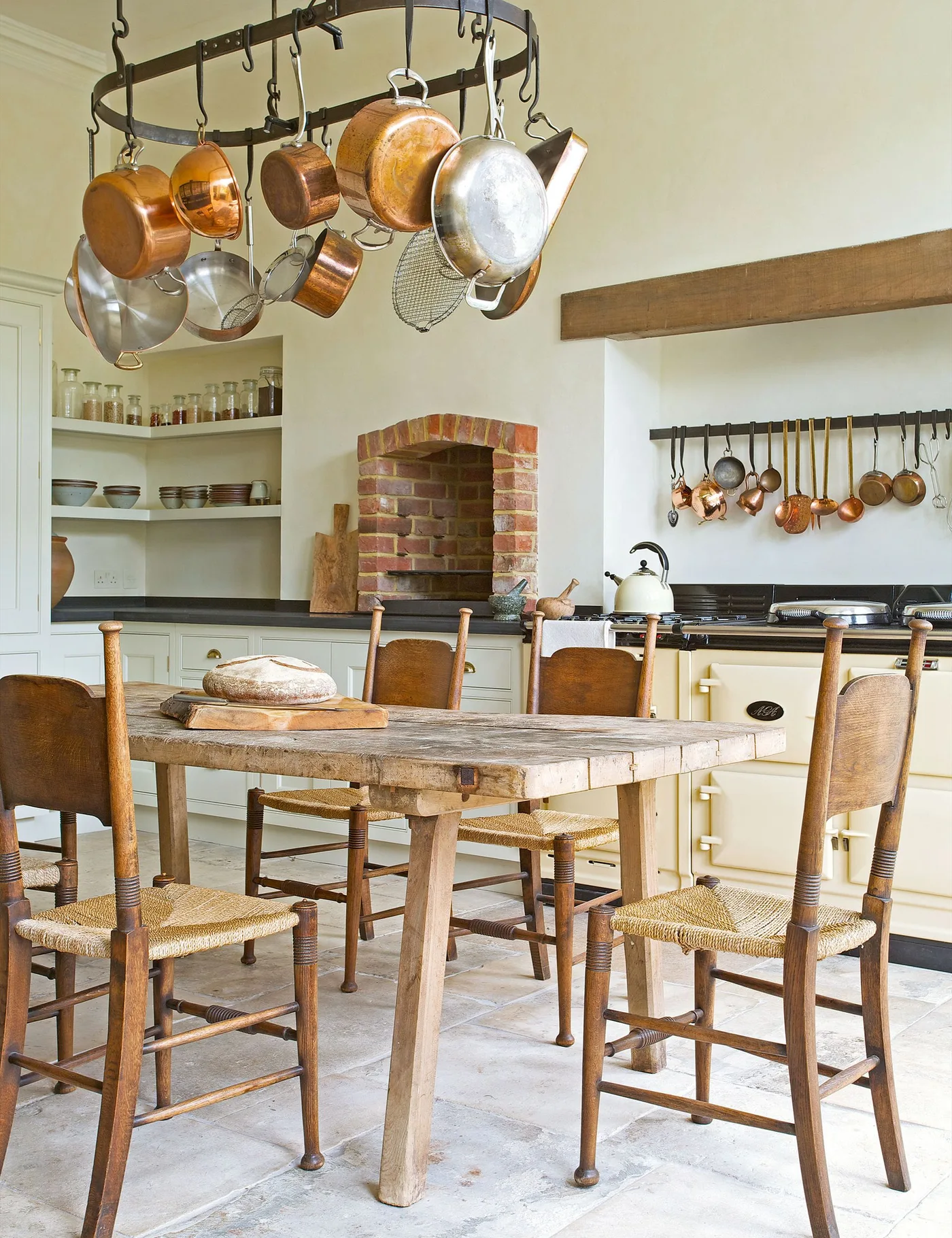 Cottage kitchen with wooden dining table and woven-seat chairs, hanging metal pans, pale cooker and open corner shelves beside a brick alcove.