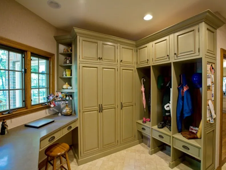 Green mudroom cabinetry with coat cubbies, drawers, bench and small desk beneath a window beside a pale floor