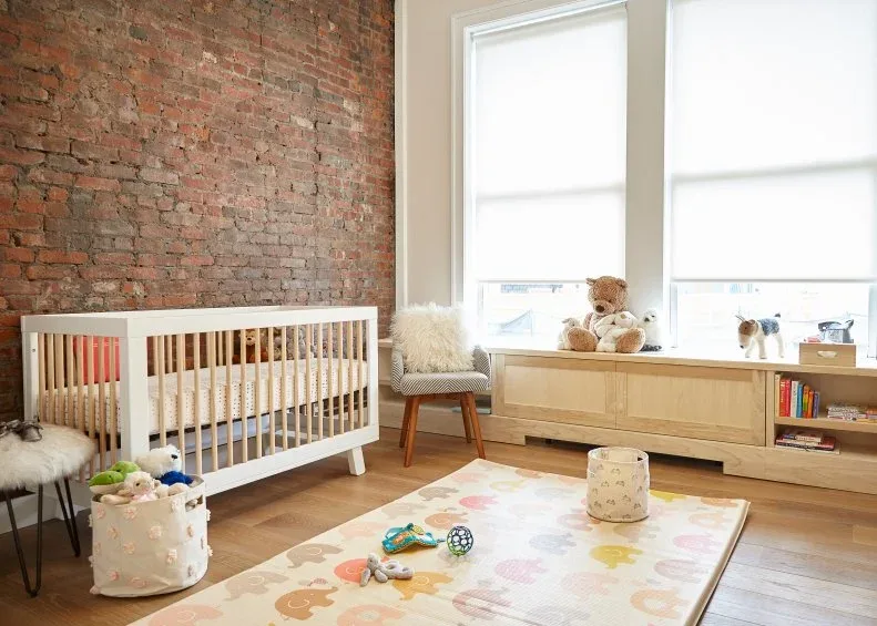 A pale wood crib and low nursery storage sit beside an exposed brick wall, broad windows, a small chair and a colorful patterned play rug