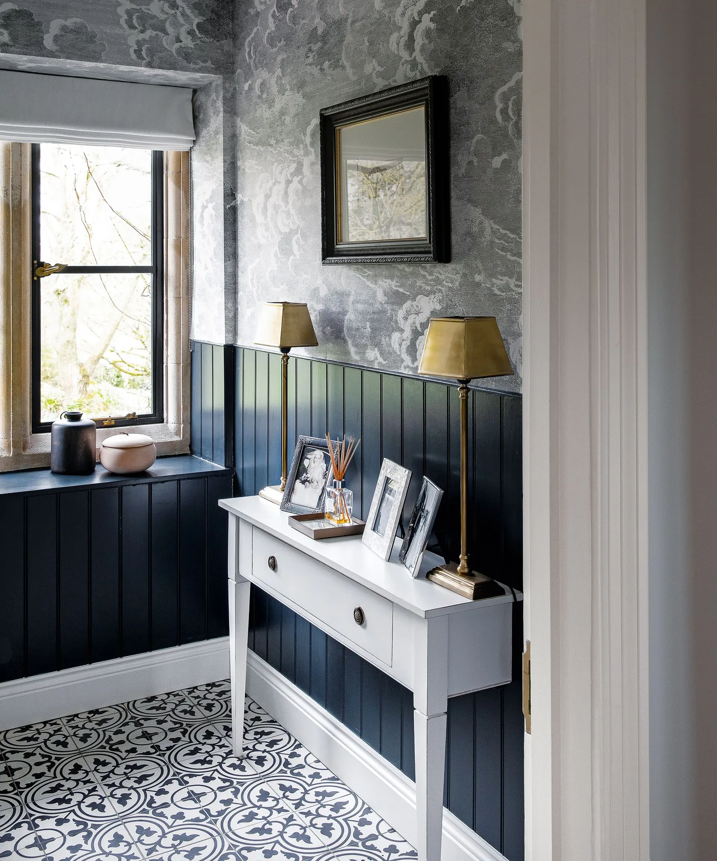 White narrow console with family frames and two gold-shaded lamps beneath blue-gray patterned wallpaper and dark paneling