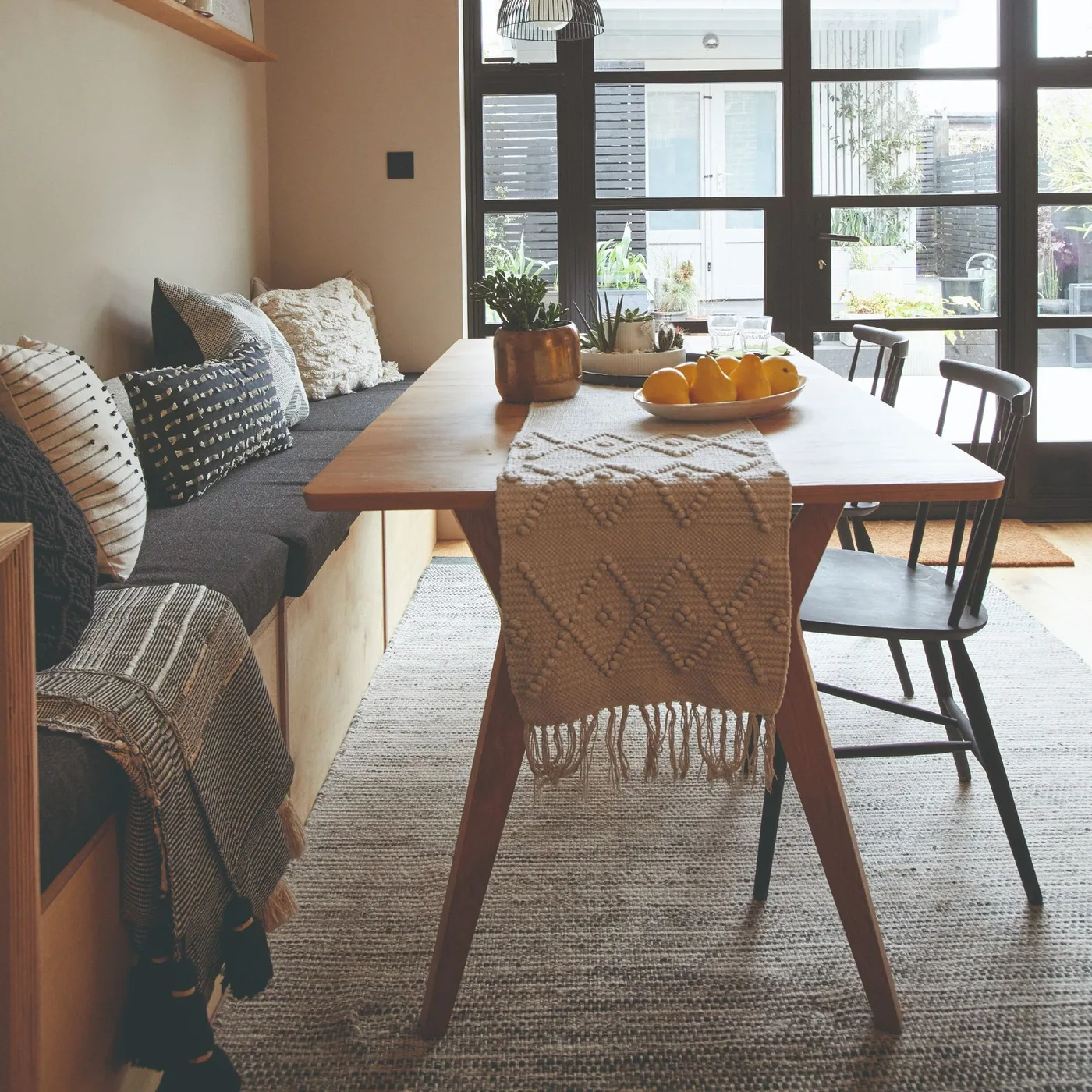 A wooden table with a textured runner stands beside a gray cushioned bench, dark spindle chairs and broad dark-framed windows