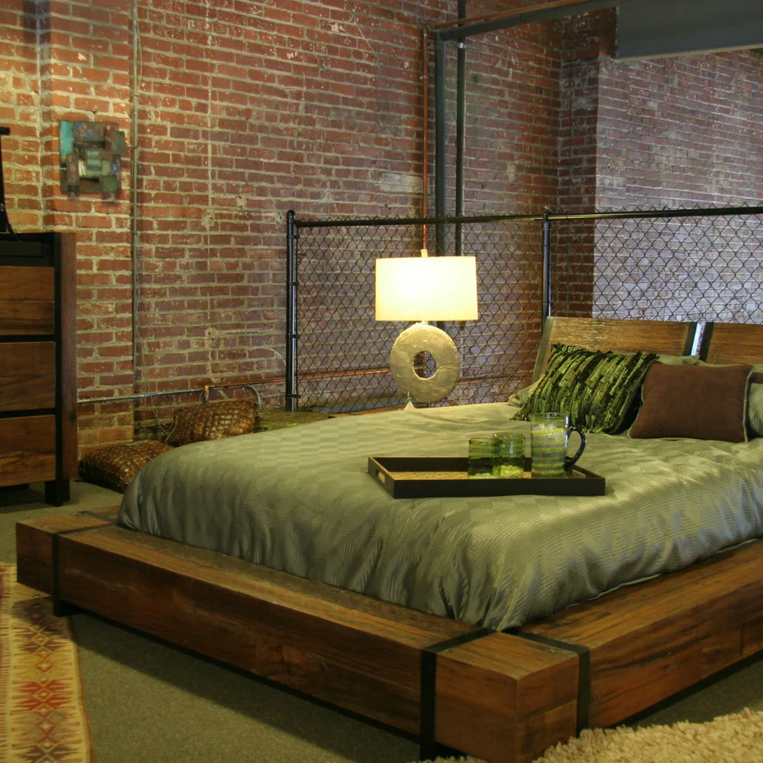 A low wooden platform bed with green bedding sits against exposed brick and a dark mesh partition beside a pale-shaded lamp