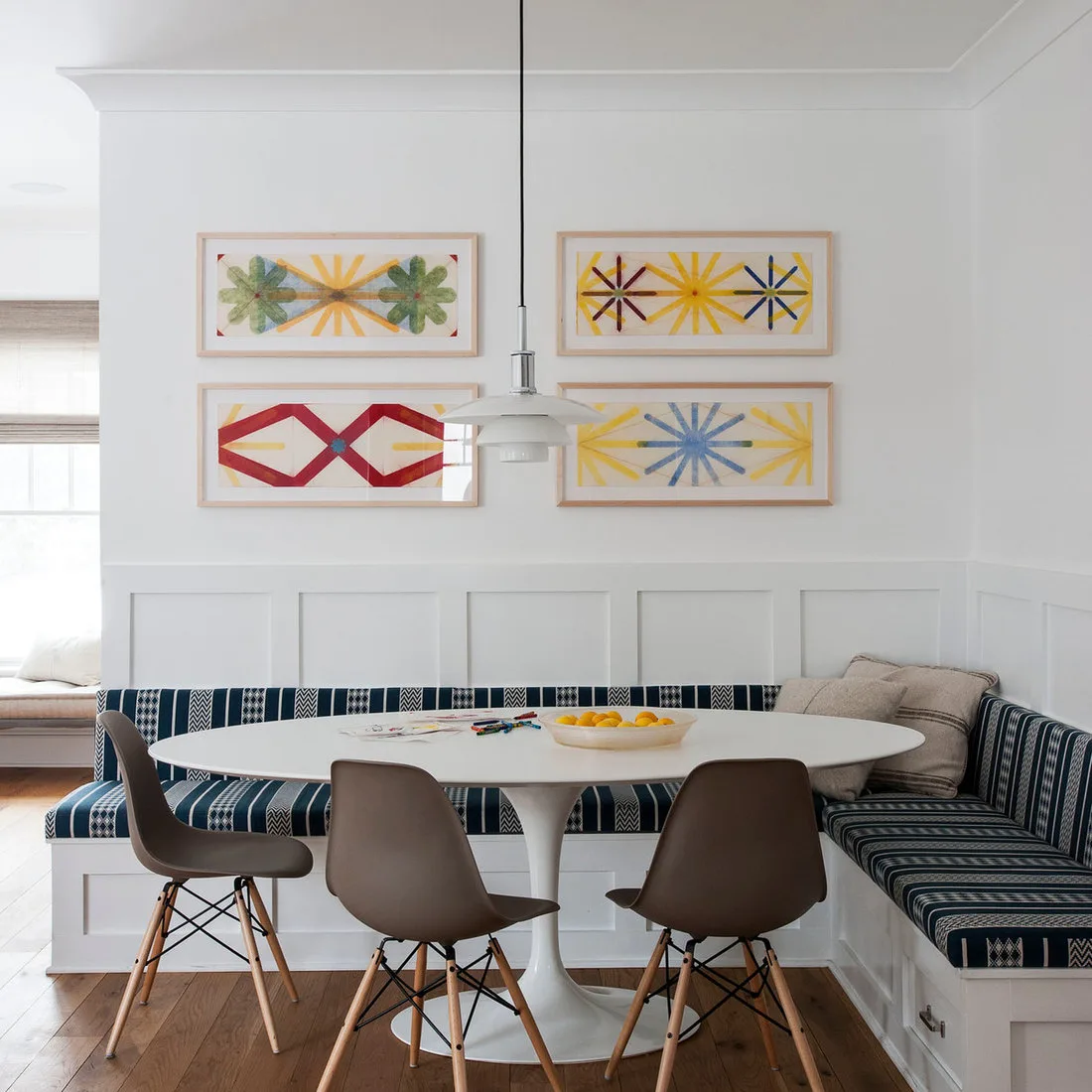 White oval pedestal table with brown chairs and a blue-striped corner banquette beneath colorful framed art