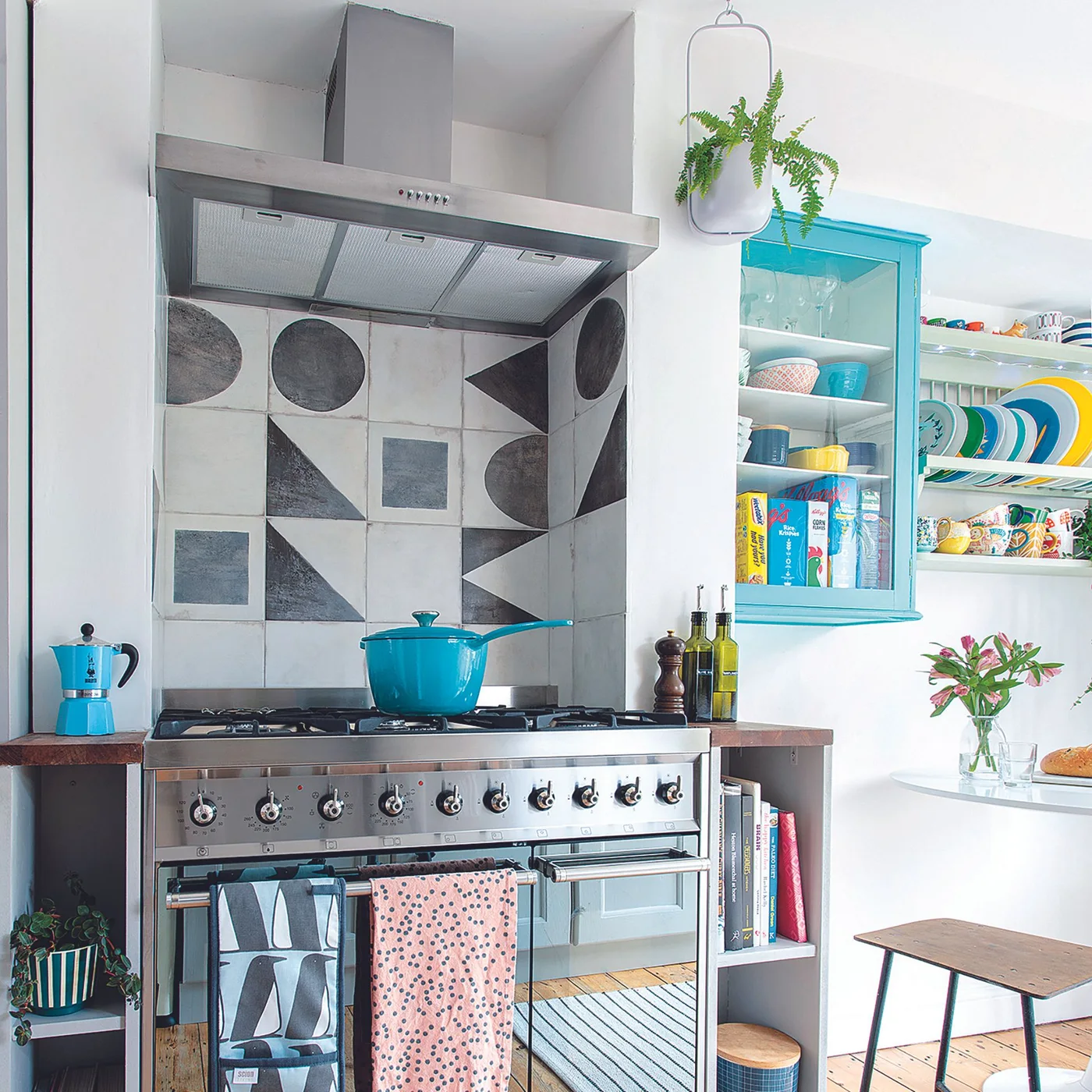 Black-and-gray geometric tiles form a patterned cooking backsplash beneath a metal hood beside turquoise cookware and shelving