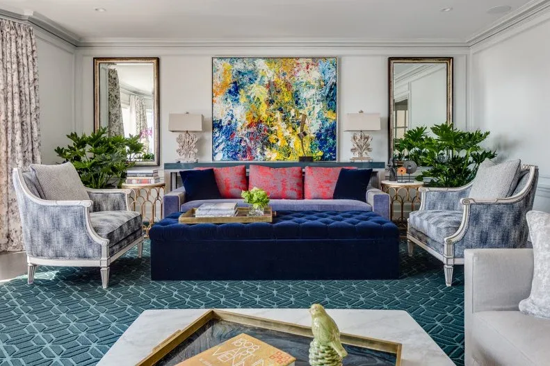 Living room with colorful abstract artwork, blue sofa and tufted ottoman, pink cushions, gray armchairs, green patterned rug and paired mirrors.