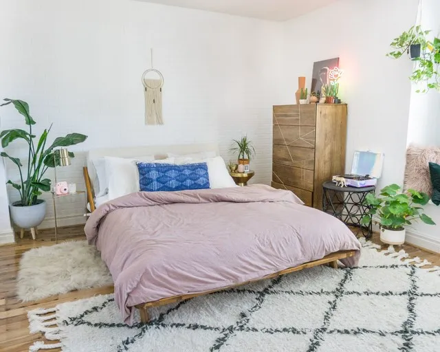 A muted pink duvet covers a low bed beside timber storage and a leafy plant.