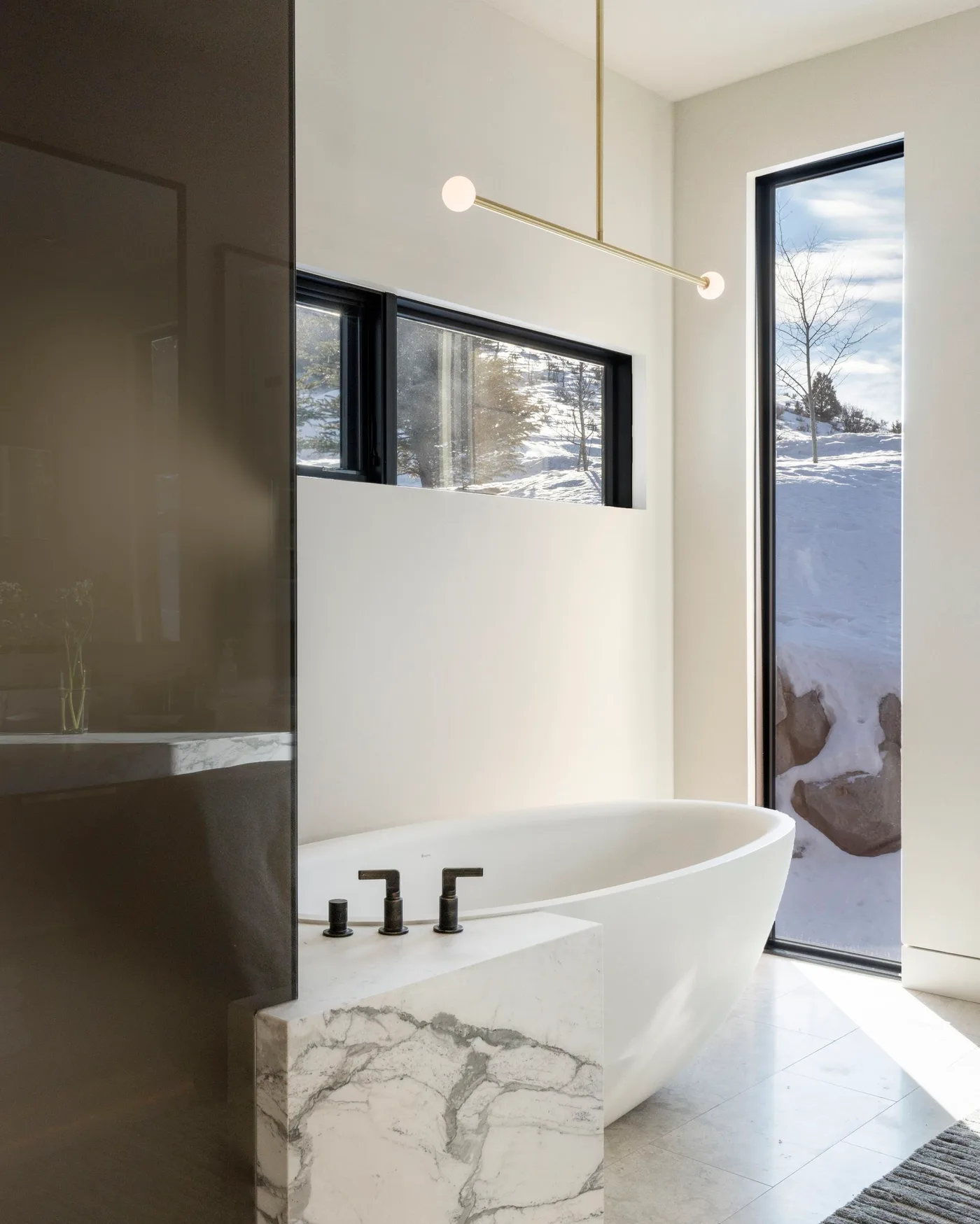 White freestanding bath beside cream walls, tall dark-framed windows and a veined front ledge
