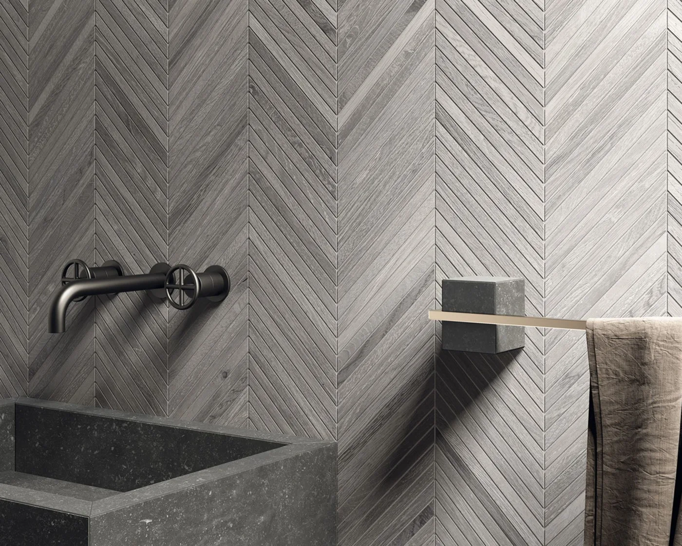 Dark gray rectangular basin beneath a black wall tap, gray narrow chevron wall tiles, and a slim towel rail with a neutral towel.