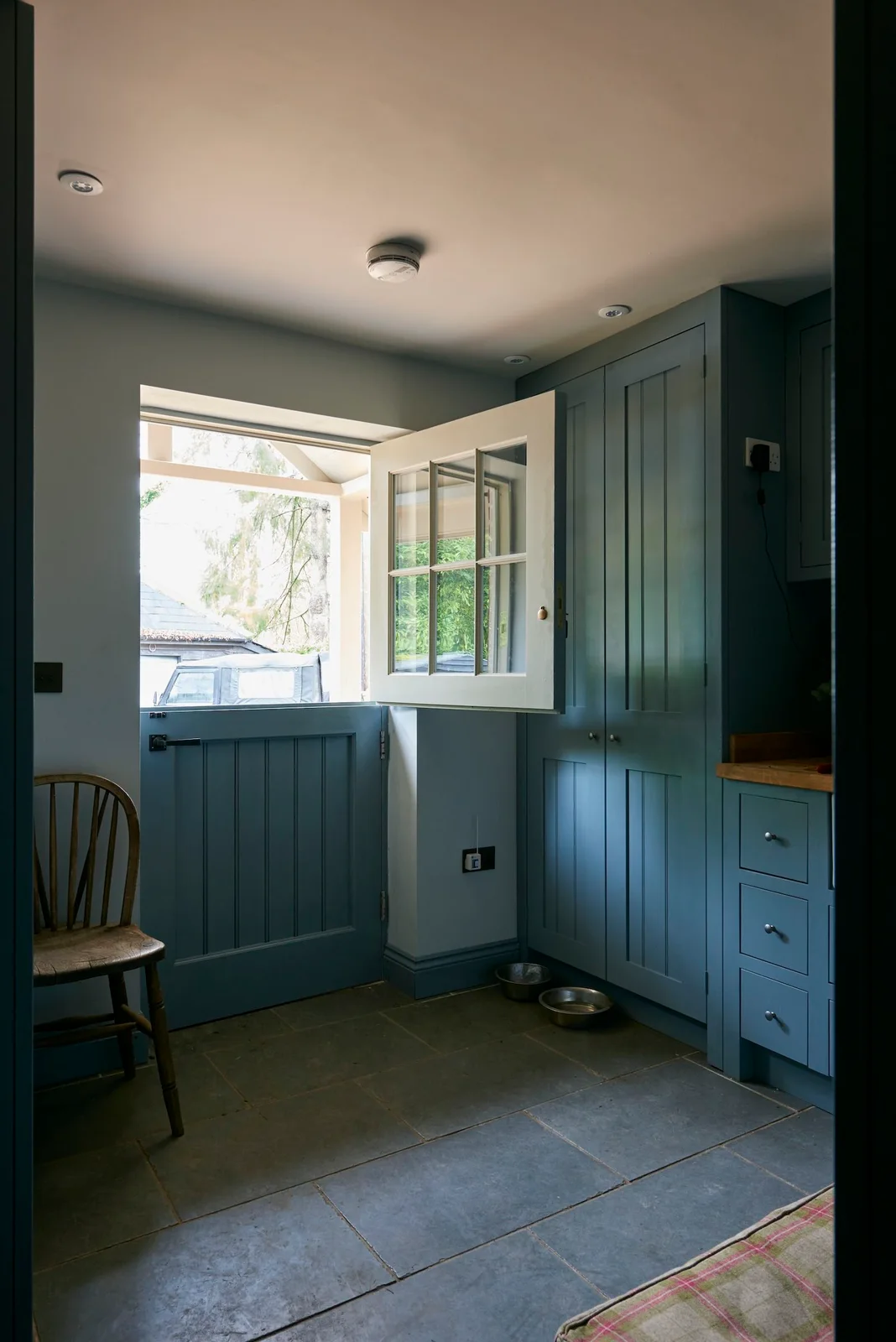 A blue divided exterior door sits beside blue framed cupboards and drawers, dark floor tiles and a simple wood chair in a compact entrance