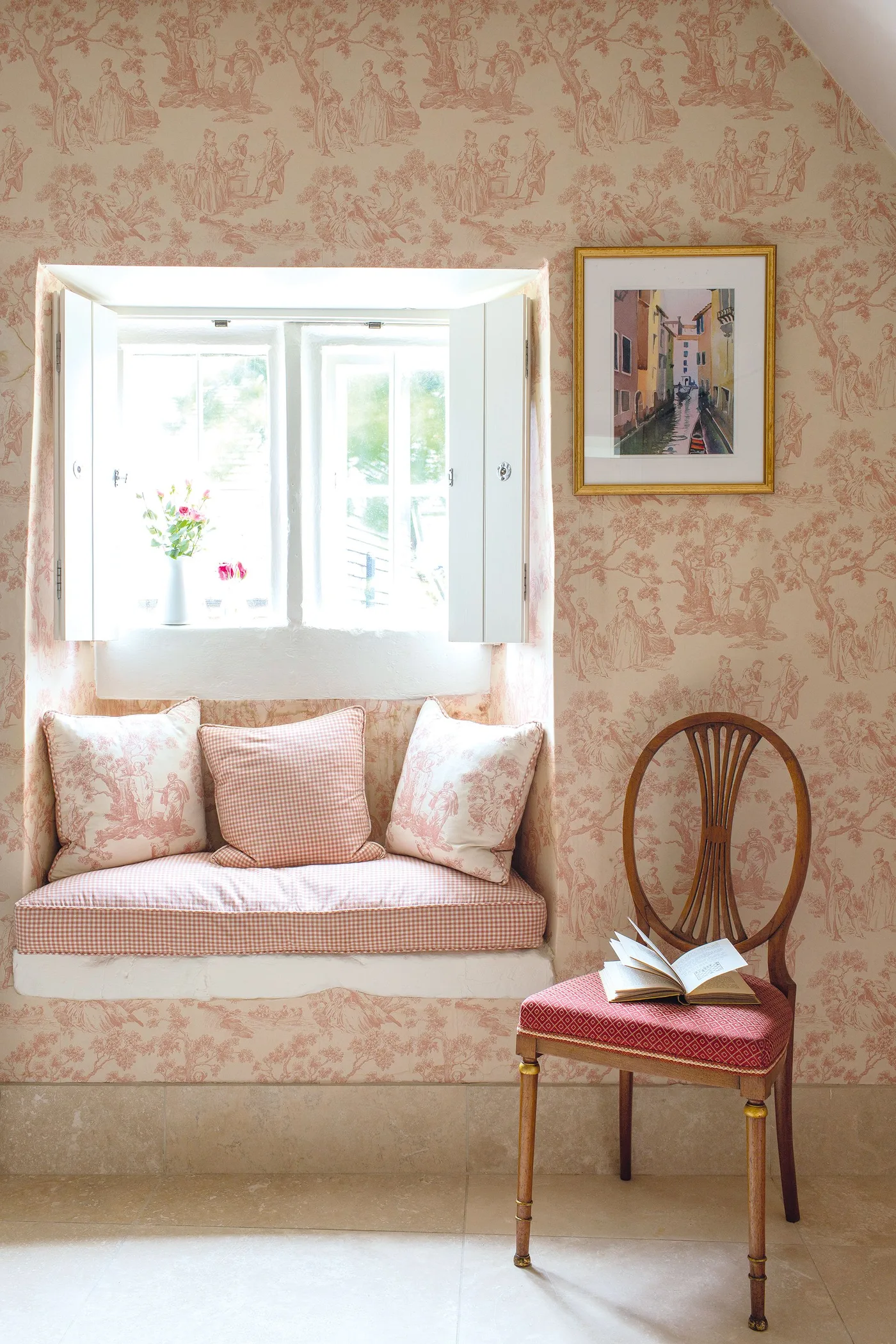 Pink patterned bedroom corner with a recessed cushioned window seat and a curved wooden chair with a red seat.