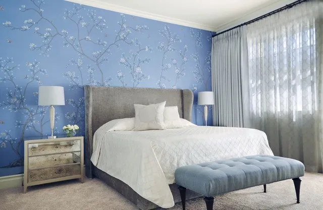 A blue botanical wall frames a gray headboard and white bedding beside pale curtains.
