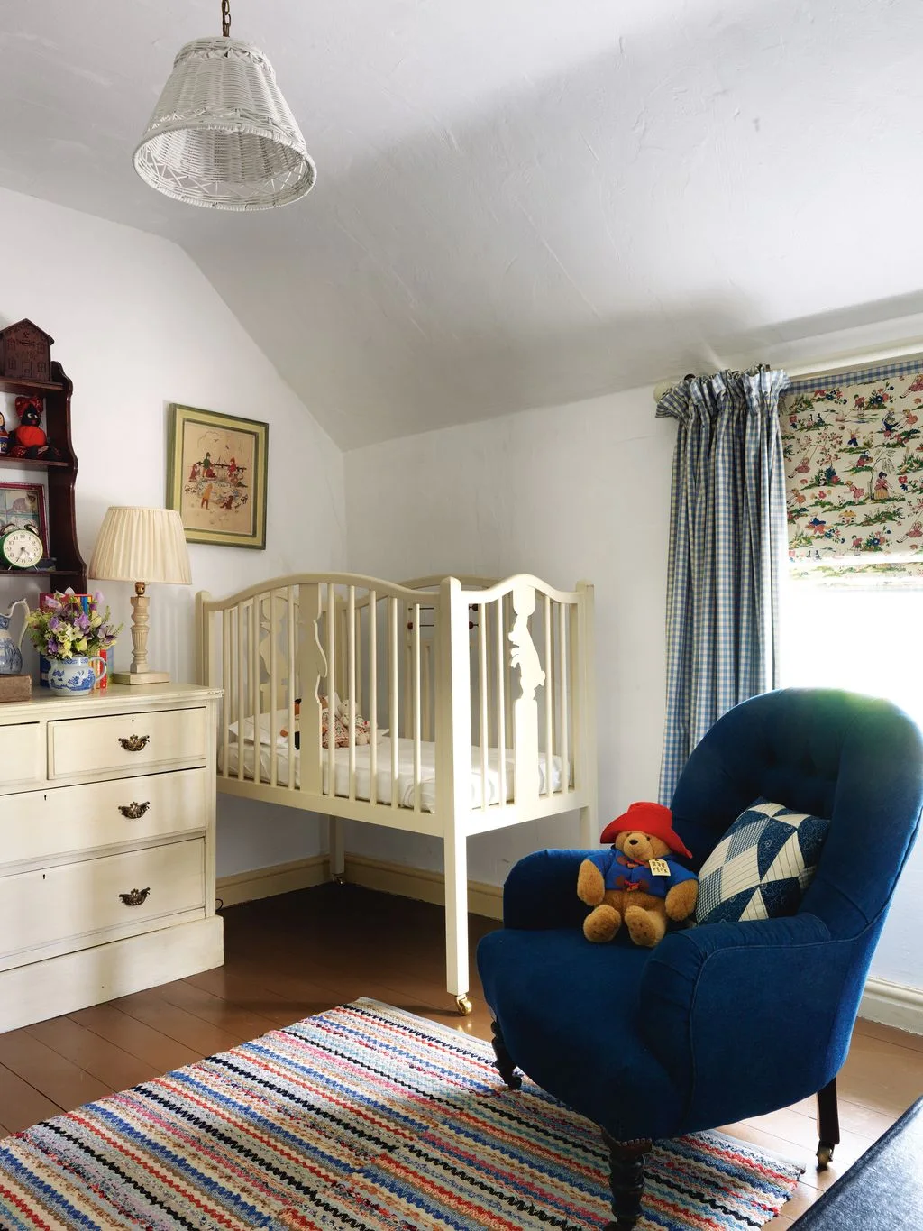 A printed nursery blind and blue checked curtains sit beside a pale crib, blue adult chair and multicolored striped rug