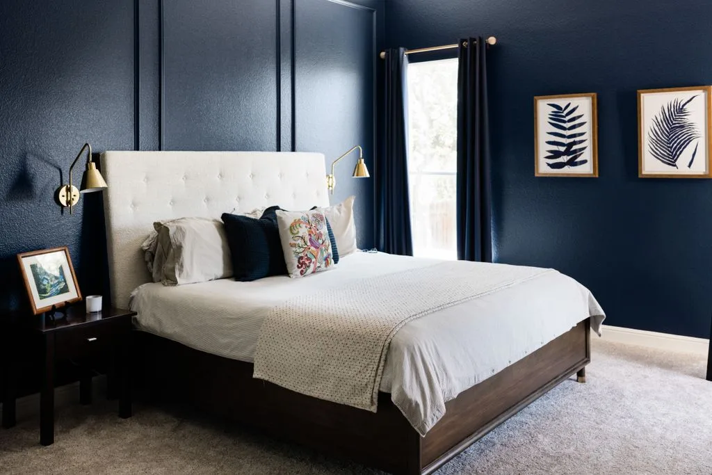Navy bedroom walls with rectangular molding, a tall cream headboard, white bedding, and gold-colored wall lights.