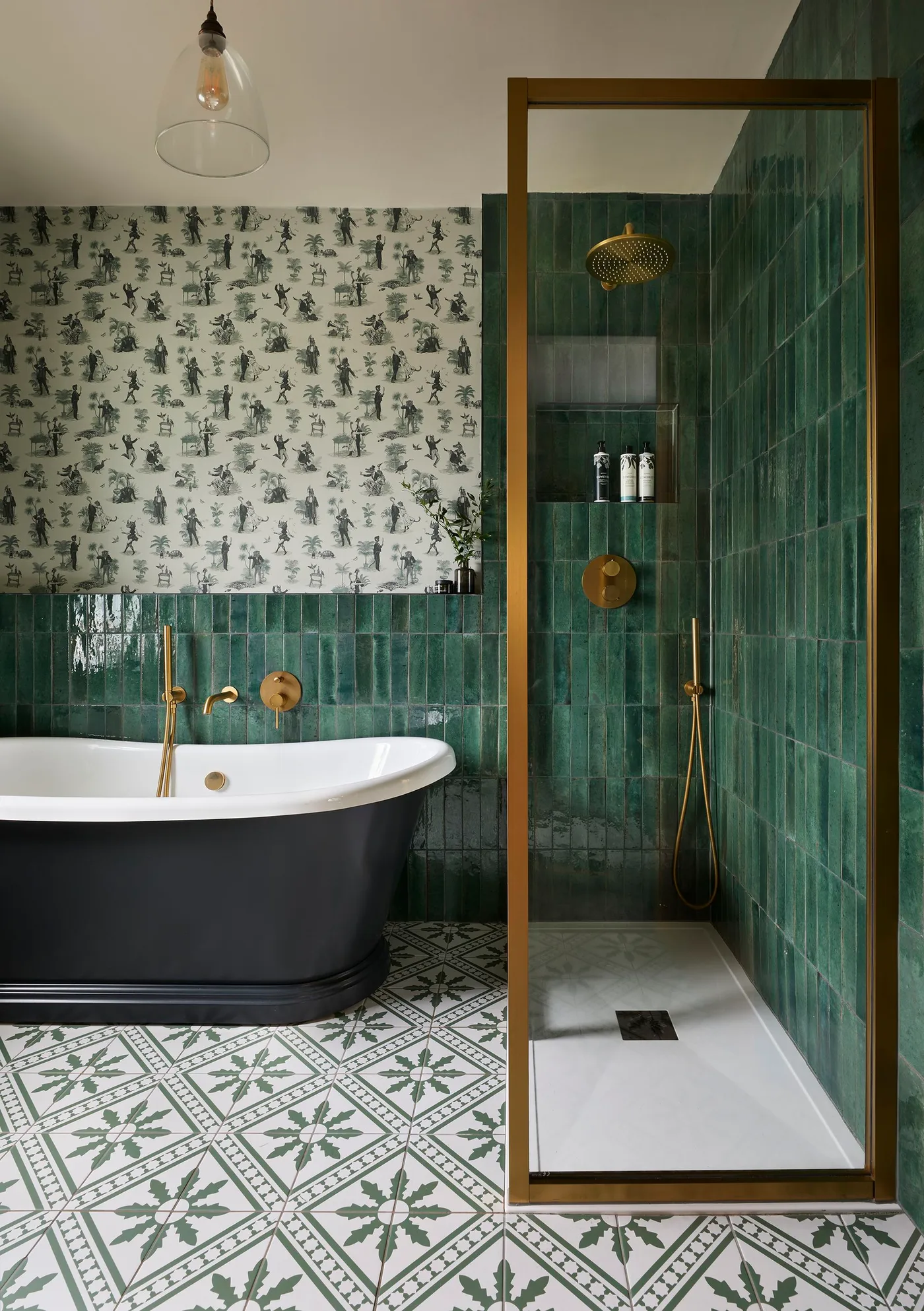 Green-tiled bathroom with a dark freestanding tub, patterned floor and warm metal-framed shower glass.