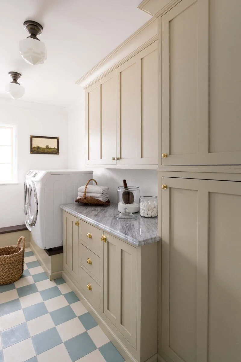 Beige laundry cupboards with pale veined counter, gold-colored knobs, blue-and-white checked floor and an appliance on a raised base