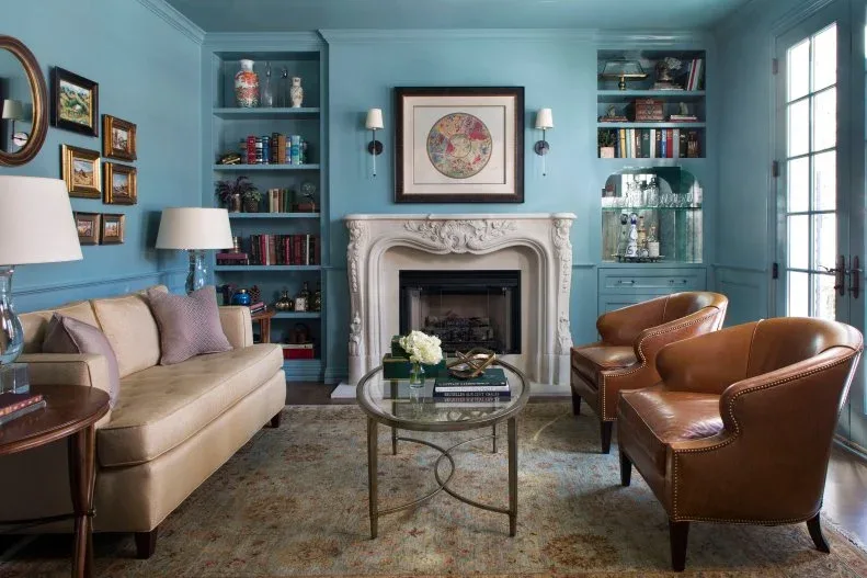 Blue paneled room with carved pale fireplace, framed artwork, built-in shelves, tan sofa and curved brown chairs