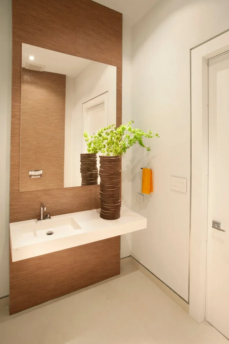 A compact white basin projects from a vertical wood-toned wall panel below a rectangular mirror beside pale walls, a glazed door and a yellow hand towel