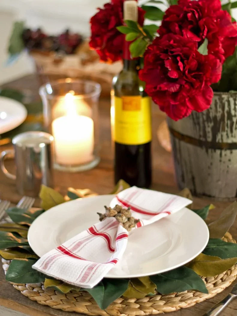 Broad green leaves sit between a woven charger and white plate with a red-striped napkin beside red flowers