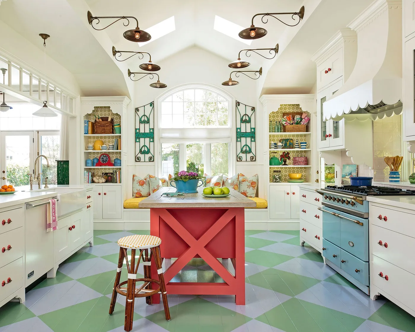 Cottage kitchen with red central island, blue cooker, pale cabinets, green-and-blue checked floor and colorful open display shelves beside an arched window.