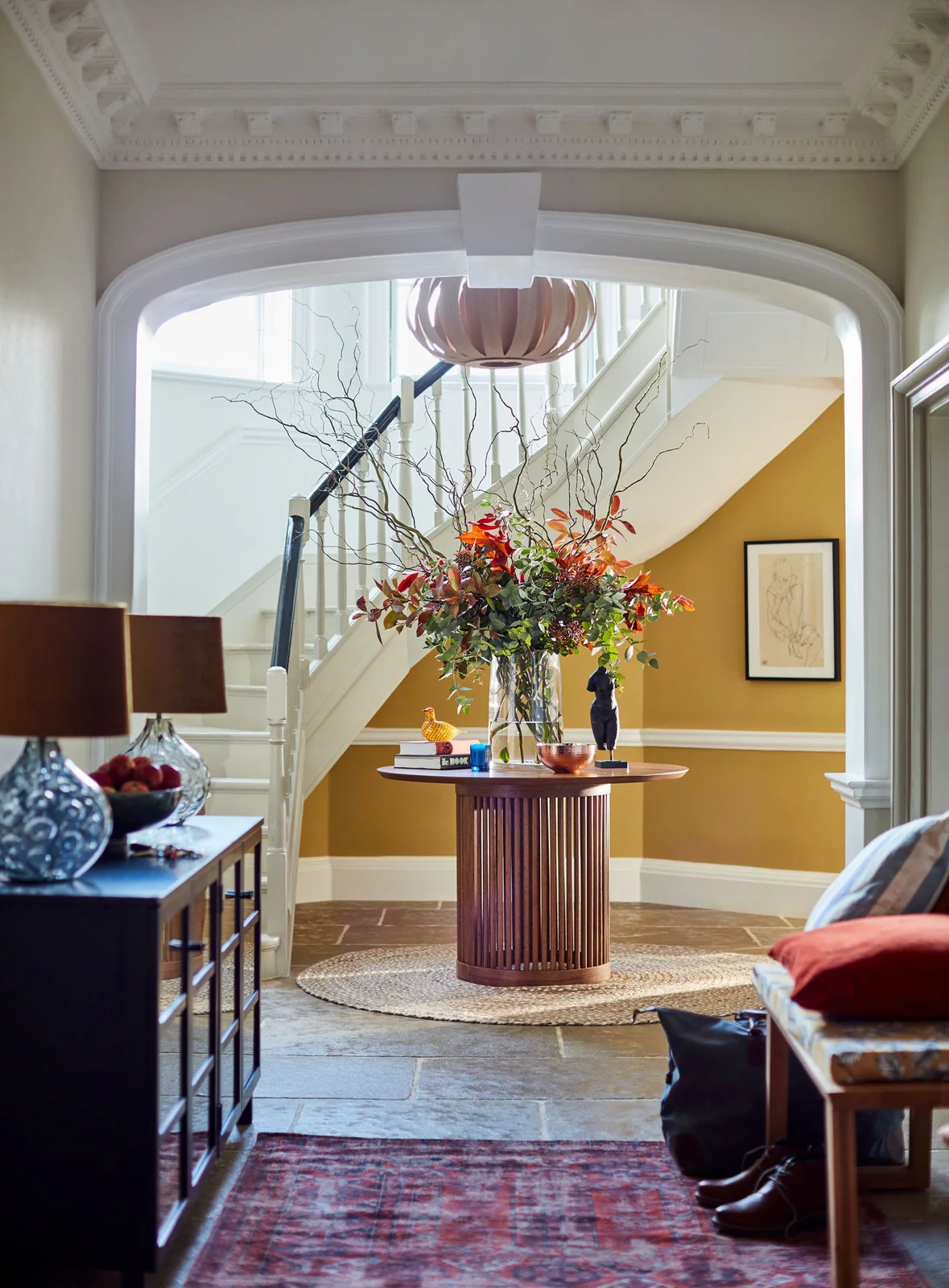 A reddish fluted round table stands beneath a pink ribbed pendant beside a mustard stair recess, blue-and-white vases and a patterned red rug
