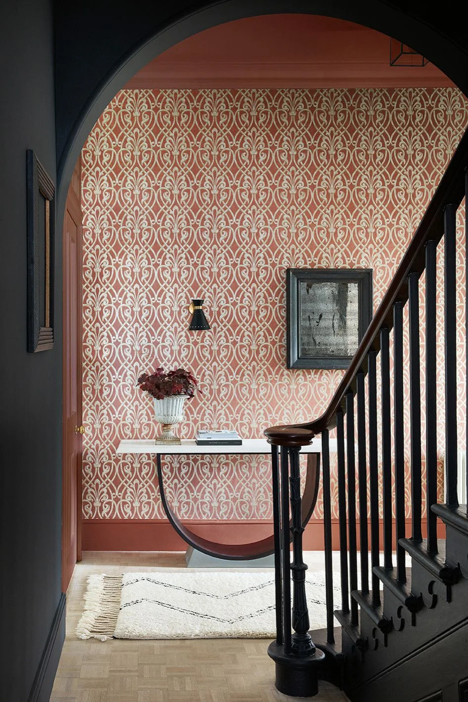 Coral-and-cream patterned wallpaper surrounds a narrow console with flowers beneath an arch beside a dark stair railing and pale rug