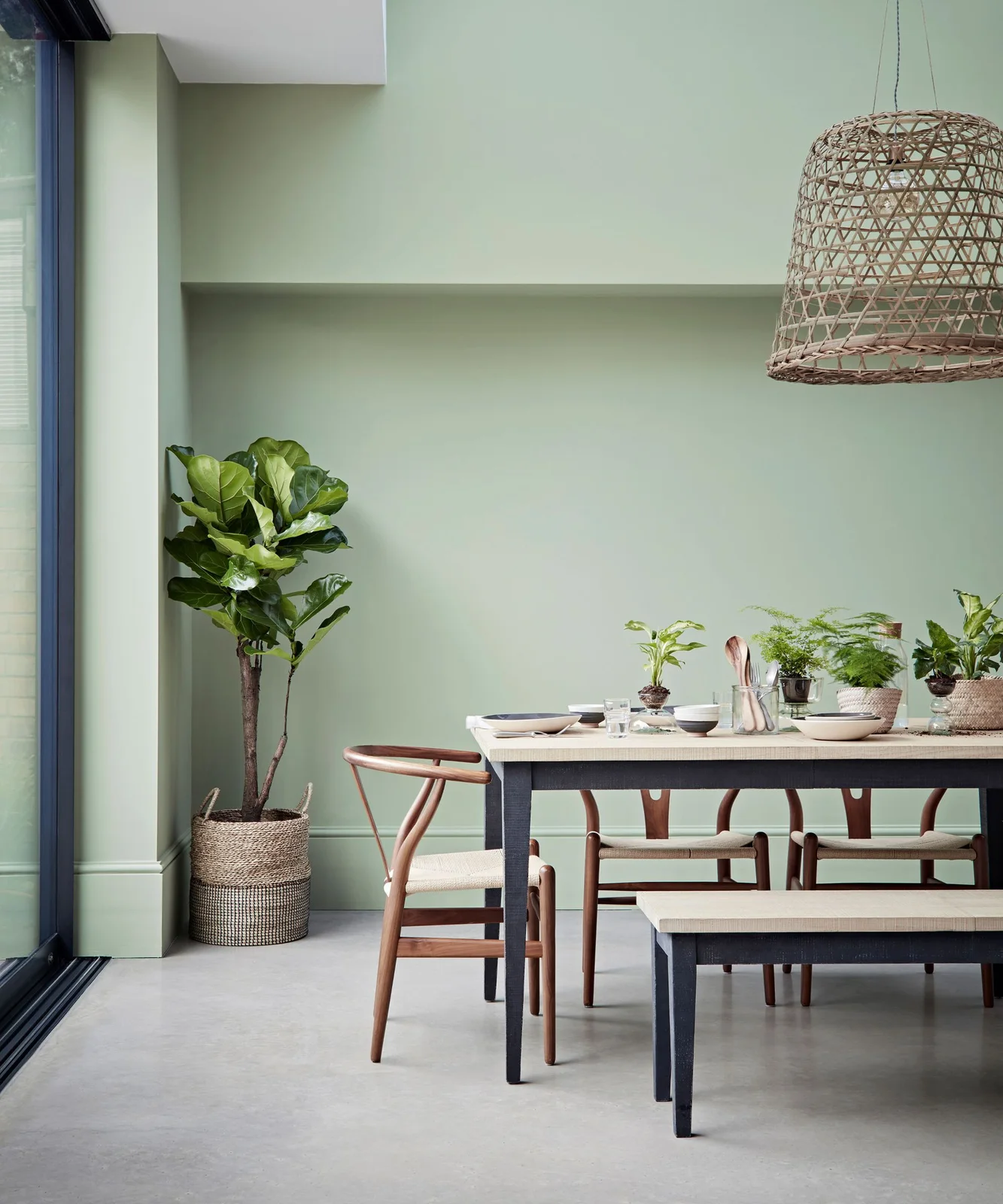 A wood-topped dining table with dark legs stands beside wooden chairs, a matching bench, pale green walls and a woven pendant