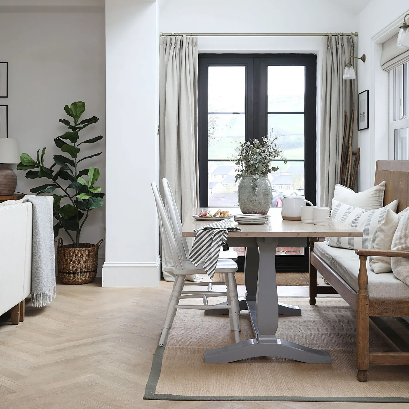 A wooden table, pale chairs and a cushioned wooden bench sit beside dark-framed glazed doors and pale curtains in a shared living space