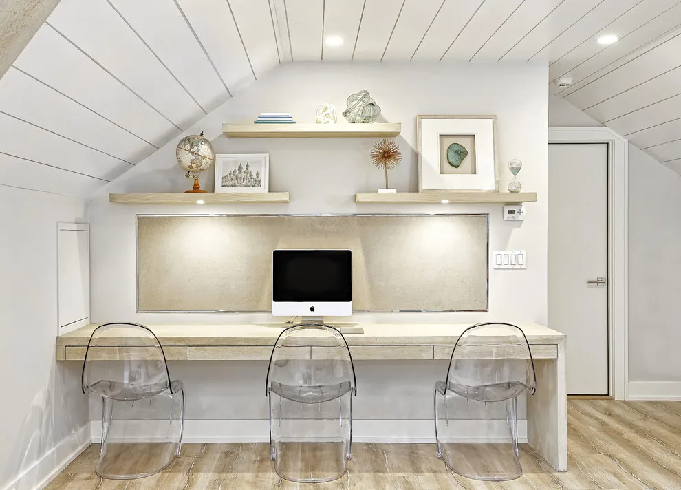 A long pale work counter with clear backed chairs sits beneath a white paneled attic ceiling