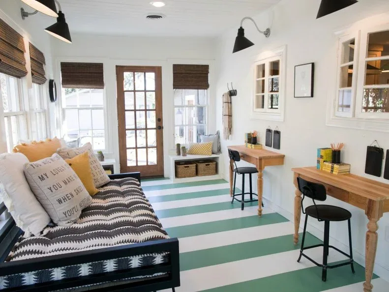 Bright country sunroom with two wood desks, dark chairs, patterned sofa, striped green-and-white floor and glazed door