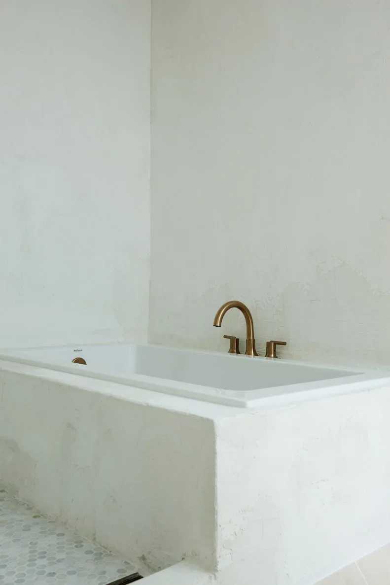 A rectangular white built-in bathtub has a broad pale surround and a gold-toned curved tap beside plain light walls and small patterned floor tiles