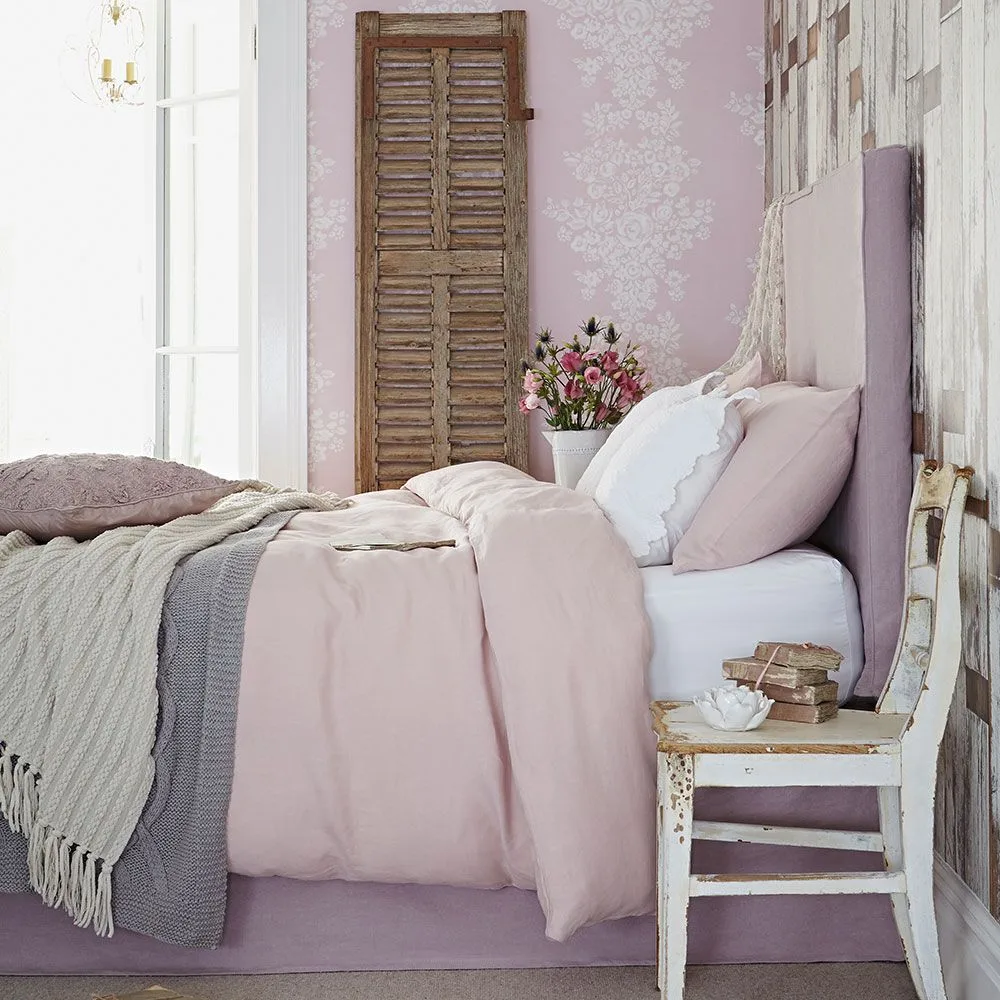 Bedroom with pink bedding and wall pattern, a tall pale pink headboard, weathered wooden chair, shutter panel and layered throws.