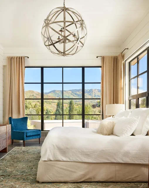A pale bed faces broad windows beneath a sculptural pendant.
